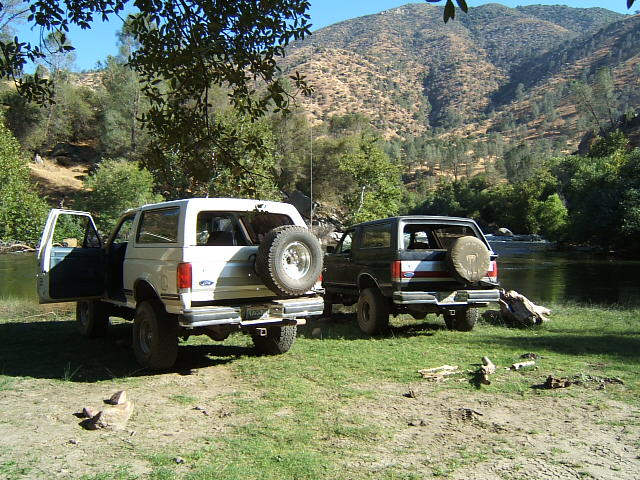 Two Ford Broncos parked on grass near a river. One is white with the driver's door open, the other is dark
