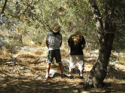 1988 Ford Bronco - Two men are urinating in a wooded area with dry brush and rocks.