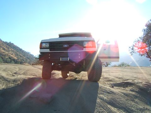 1988 Ford Bronco - A Ford Bronco sits on a dusty, rocky terrain under a bright sun, its driver's side door open.