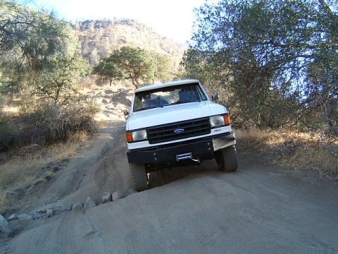 1988 Ford Bronco - White Ford truck navigates a dusty, off-road trail with arid hills in the background.