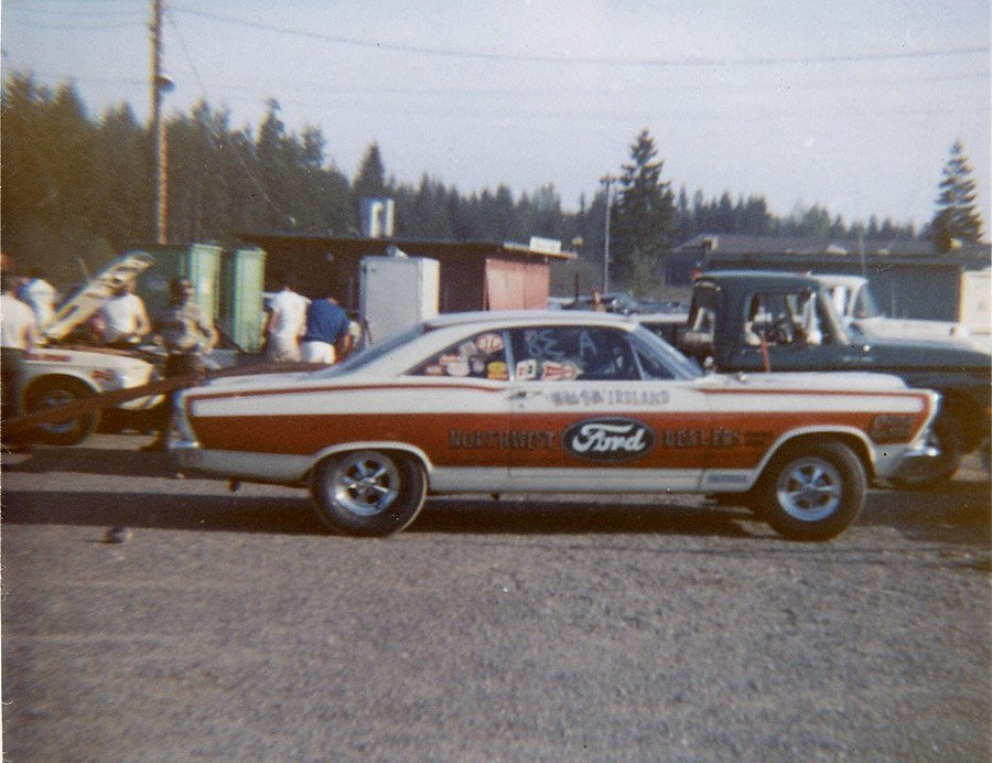 White and orange Ford drag coupe in side view at a race pit area.