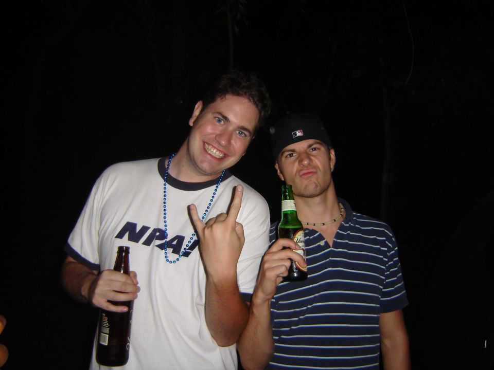 Guy in white shirt with blue beads and guy in striped shirt with black baseball cap holding beer bottles