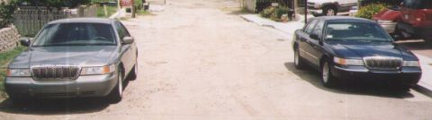 2000 Mercury Grand Marquis - Two 2000 Mercury Grand Marquis sedans are parked on a dirt road in a residential area.