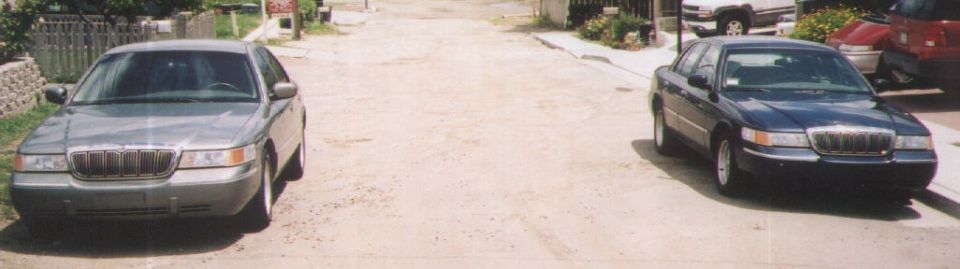 Two 2000 Mercury Grand Marquis sedans parked on a dirt road, one gray and one dark blue, viewed from the front