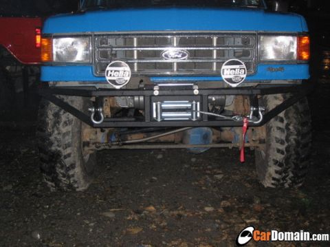 1988 Ford Bronco - Head-on night shot of a lifted blue Ford Bronco showing the chrome grille with blue Ford oval, amber turn signals, and a pair of round Hell…