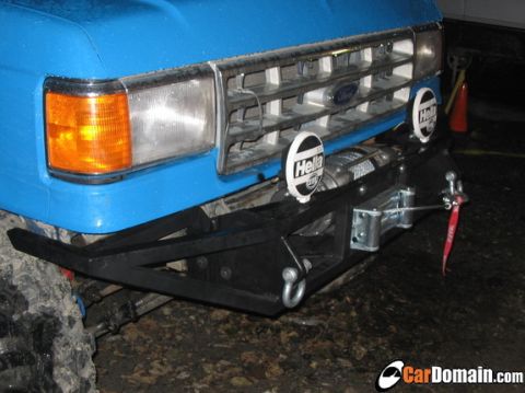 1988 Ford Bronco - Angled night close-up of the front end of a blue Ford truck, showing the chrome grille with Ford oval, amber wraparound turn signal, and co…