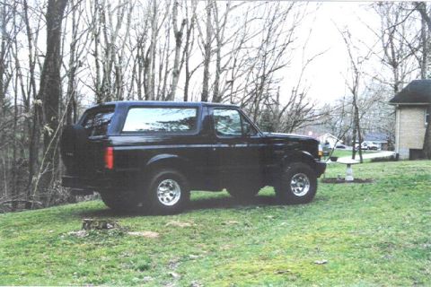 1992 Ford Bronco - This black 1992 Ford Bronco is parked on a grassy incline, with a residential house and trees visible in the background.
