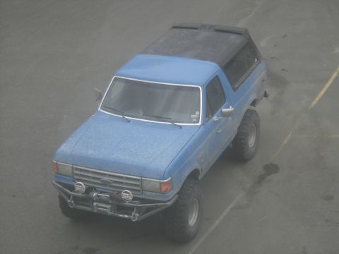 1988 Ford Bronco - Elevated three-quarter front view of a lifted blue Ford Bronco with a black removable hardtop, parked on wet asphalt and speckled with drie…