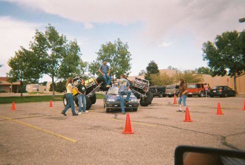 1972 Ford Ranchero - Several monster trucks are positioned on top of smaller vehicles in a parking lot, with onlookers and traffic cones present.