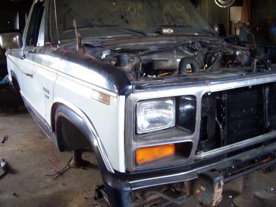 White and blue Ford Bronco with hood open, showing engine compartment. Front end is partially disassembled