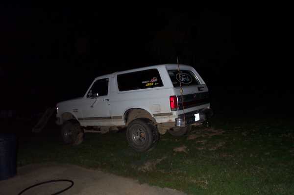 White lifted Ford Bronco at night with mud on the body and tall whip antenna, rear three-quarter view