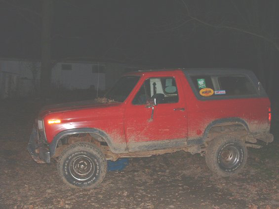 Muddy red lifted Ford Bronco side view at night on white-letter all-terrain tires
