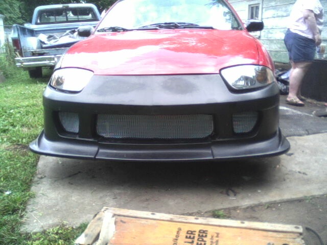 Front view of a red car with a black aftermarket bumper and mesh grille. A blue pickup truck is visible in the background