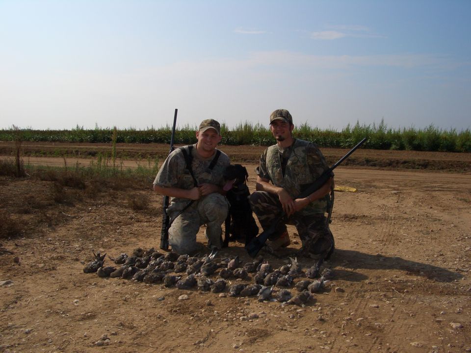 Two men in camouflage, a black dog, and a pile of birds on a dirt road
