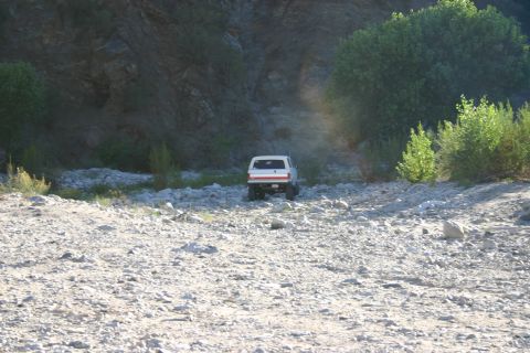 1990 Ford Bronco - distant white SUV drives away across a rocky dry riverbed beneath a steep rock face and green brush.