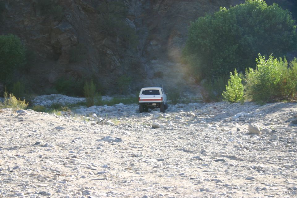 Distant white SUV driving across a rocky dry riverbed below a steep rock face