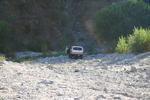 1990 Ford Bronco - white SUV with its driver door open sits in a rocky dry riverbed below a steep rock wall and brush.