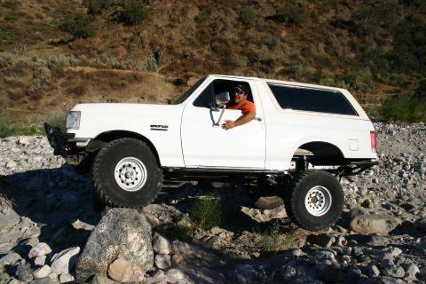 1990 Ford Bronco - lifted white Ford Bronco on large off-road tires crawls over rocks in a riverbed as the driver leans out the window.