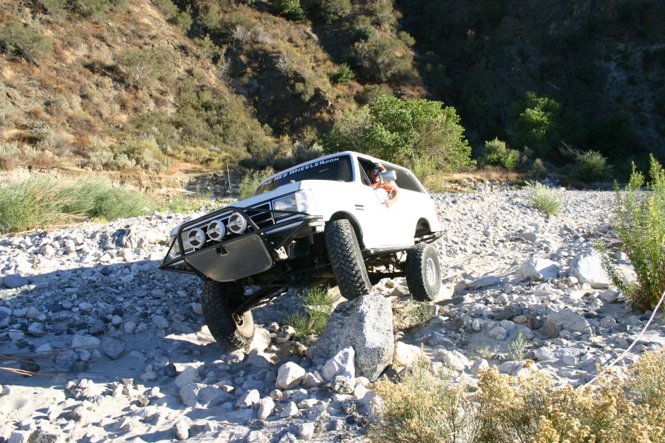 White off-road SUV with tube bumper and lights crawling over large rocks in a boulder-strewn riverbed
