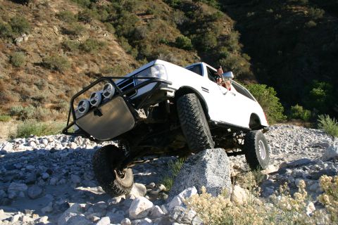 1990 Ford Bronco - white lifted SUV with a brush guard and three driving lights perches atop boulders in a rocky, brush-lined canyon wash.