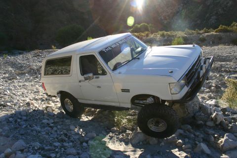 1990 Ford Bronco - white Ford Bronco lettered with a 'The Web Wheeler' windshield banner sits angled on a rocky wash with sun flare overhead.