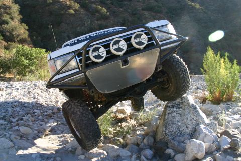 1990 Ford Bronco - low front view of a white lifted SUV with a tube brush guard and three driving lights balancing one tire atop a tall rock.