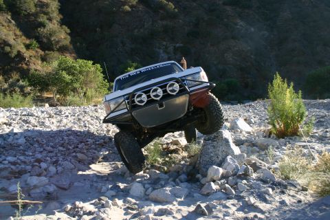 1990 Ford Bronco - white lifted SUV with a brush guard and three round driving lights crawls head-on over loose rocks in a sunlit canyon wash.