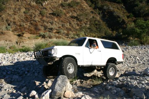 1990 Ford Bronco - white lifted Ford Bronco on chunky tires and a tubular front bumper sits among rocks in a dry riverbed below a brushy slope.