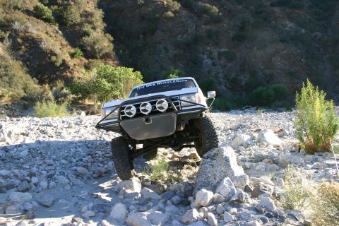 1990 Ford Bronco - front of a white lifted SUV displaying a 'The Web Wheeler' windshield banner and three driving lights as it climbs rocks.