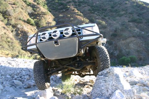1990 Ford Bronco - low front angle of a lifted SUV with a black brush guard and three driving lights articulating its suspension over rocks.