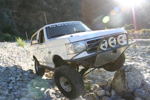 1990 Ford Bronco - white lifted Ford Bronco with a 'The Web Wheeler' banner and three driving lights sits on rocks with bright sun flare behind.