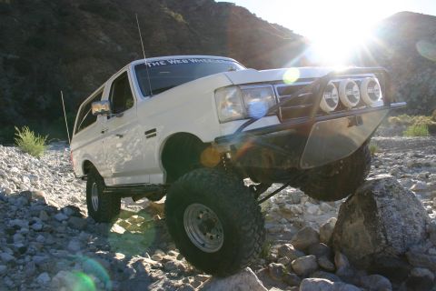 1990 Ford Bronco - white lifted Ford Bronco with a brush guard and three driving lights balances on a rock as the sun bursts over the canyon rim.