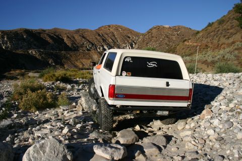 1990 Ford Bronco - rear of a white Ford Bronco with a red tailgate stripe and visible license plate sits on rocks in a dry canyon under blue sky.