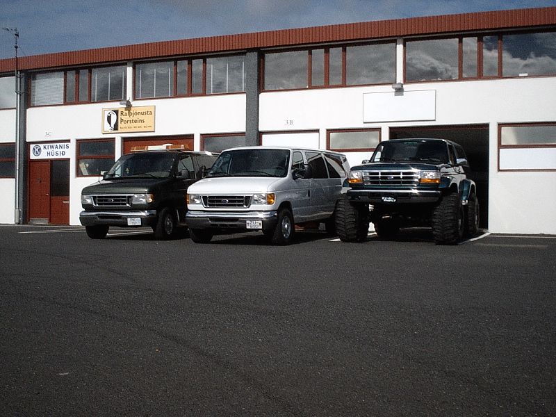 Lifted dark Ford Bronco on big tires beside a silver van and green vehicle in a paved lot