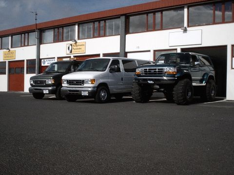 1996 Ford Bronco - A lifted dark green 1996 Ford Bronco with oversized tires is parked alongside two vans in front of a commercial building.