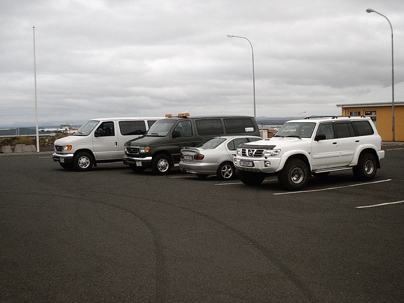 White van, gray van, silver sedan, and white SUV parked in an overcast lot.