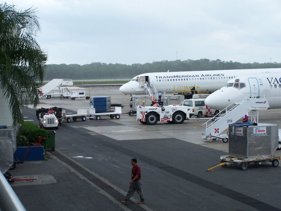Airport tarmac with Transmeridian Airlines plane, baggage carts, and tugs under an overcast sky