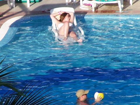 1979 Ford F-150 - A woman reclines in a pool chair while a man holds a ball in the water, with palm fronds in the foreground.