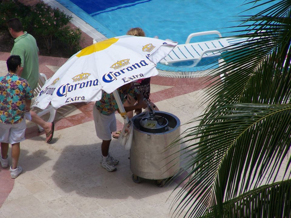 People stand near a pool under a large Corona umbrella. A cooler is next to them