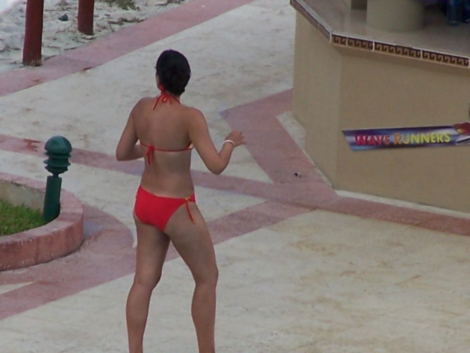 Woman in red bikini from behind, standing on a paved area with sand in the background. A sign reads WAVE RUNNERS