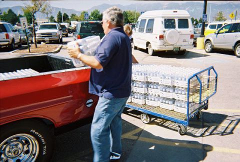 1972 Ford Ranchero - A man loads cases of bottled water into the bed of a red pickup truck in a parking lot.