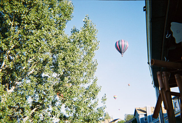 hot air balloons in sky over houses, tree on left, part of building on right
