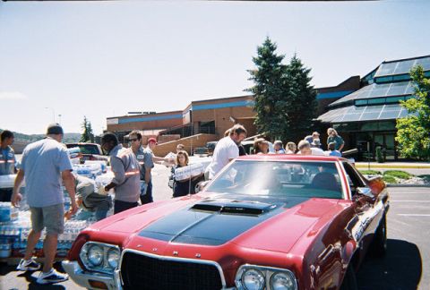1972 Ford Ranchero - A red 1972 Ford Ranchero with a black hood scoop and racing stripes is parked outside a building while people gather around.