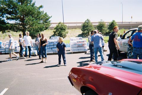 1972 Ford Ranchero - A red car's front end is visible in the foreground, with a group of people and stacked water bottles in the background.