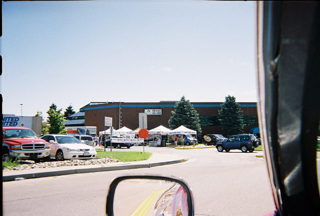 View past red fender and side mirror toward white vendor canopies at a swap meet parking lot