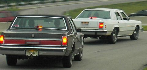 1985 Ford LTD Crown Victoria - Two sedans drive on a road, one dark and one white, with the dark car in the foreground.