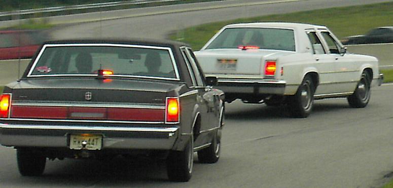 Dark sedan with red stripe and chrome trim, followed by a white sedan. Both have their brake lights on