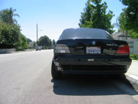 1985 Ford Bronco - A black BMW 740iL sedan is parked on the side of a street, showing damage to its rear bumper and taillights.