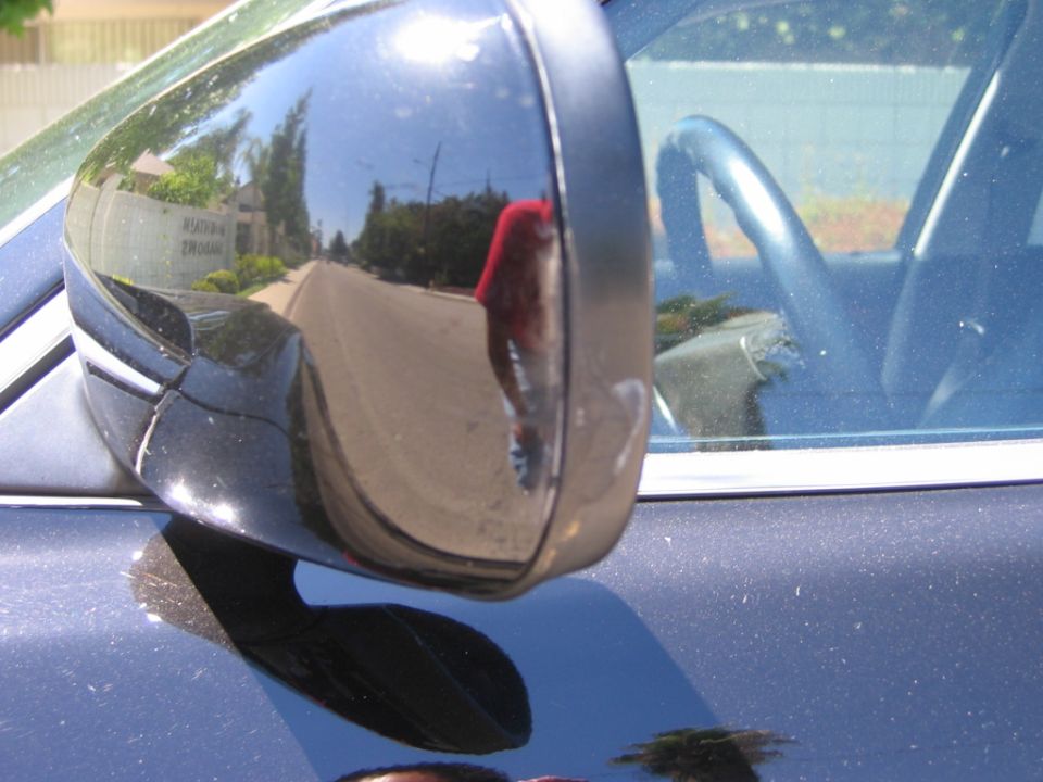 Close-up of a glossy dark blue side mirror housing reflecting the street, steering wheel visible through the glass