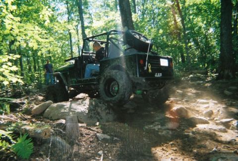 1976 GMC C/K Pickup - An off-road vehicle tackles a challenging rocky path in a wooded area.